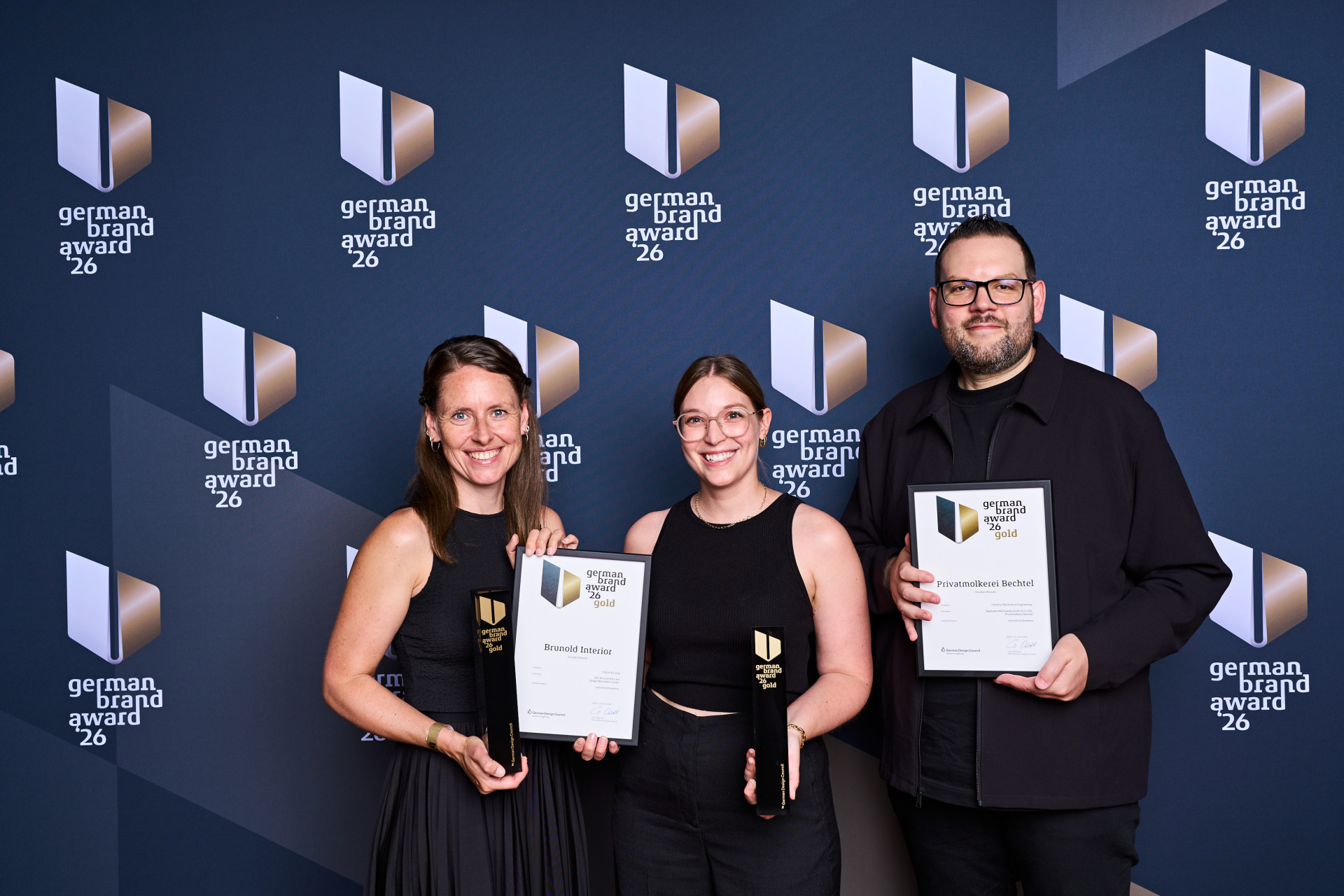 Drei Personen halten Zertifikate und Trophäen beim German Brand Award. Der Hintergrund zeigt das Logo des Awards. Alle lächeln in die Kamera.