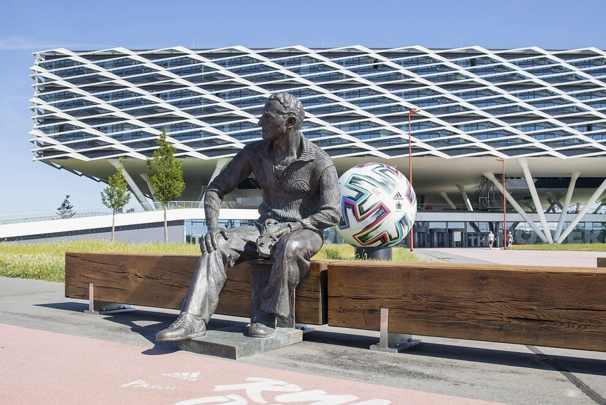 Eine Statue eines sitzenden Mannes vor einem modernen Gebäude. Daneben ein großer Fußball. Im Hintergrund ist ein architektonisch auffälliges Gebäude zu sehen.