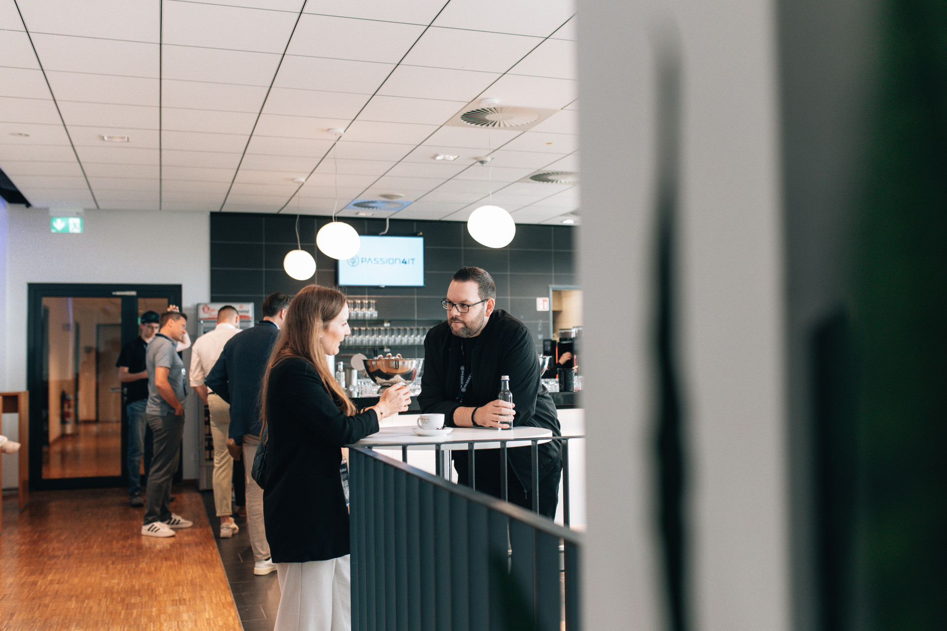Zwei Personen unterhalten sich an einem Stehtisch in einem modernen Büro oder Café. Im Hintergrund sind weitere Personen und eine Kaffeebar zu sehen.