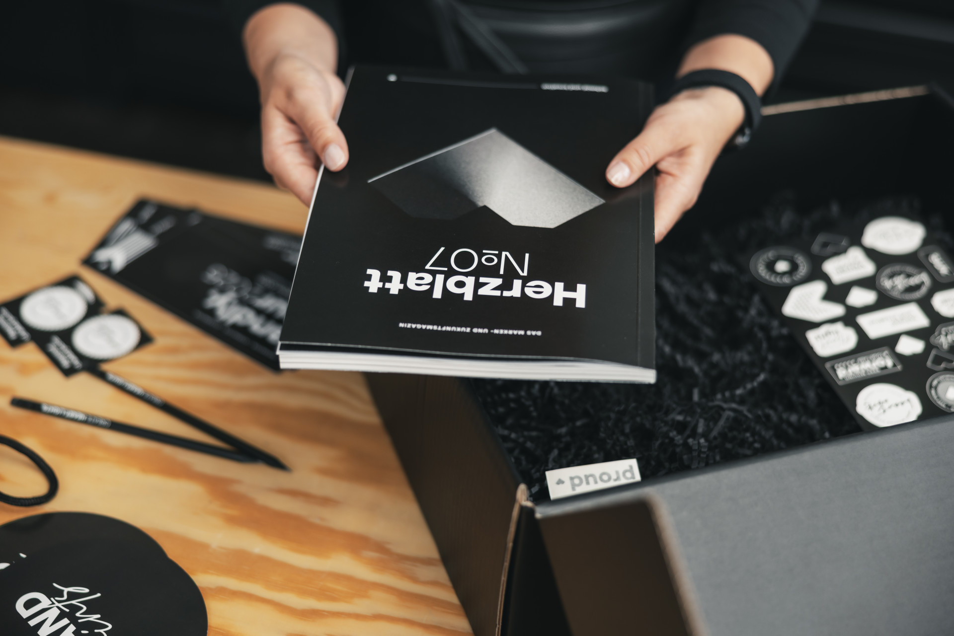 Eine Person hält ein schwarz-weißes Magazin mit der Aufschrift „Herzblätt No. 7“. Im Hintergrund sind einige Aufkleber und Stifte auf einem Holztisch zu sehen.