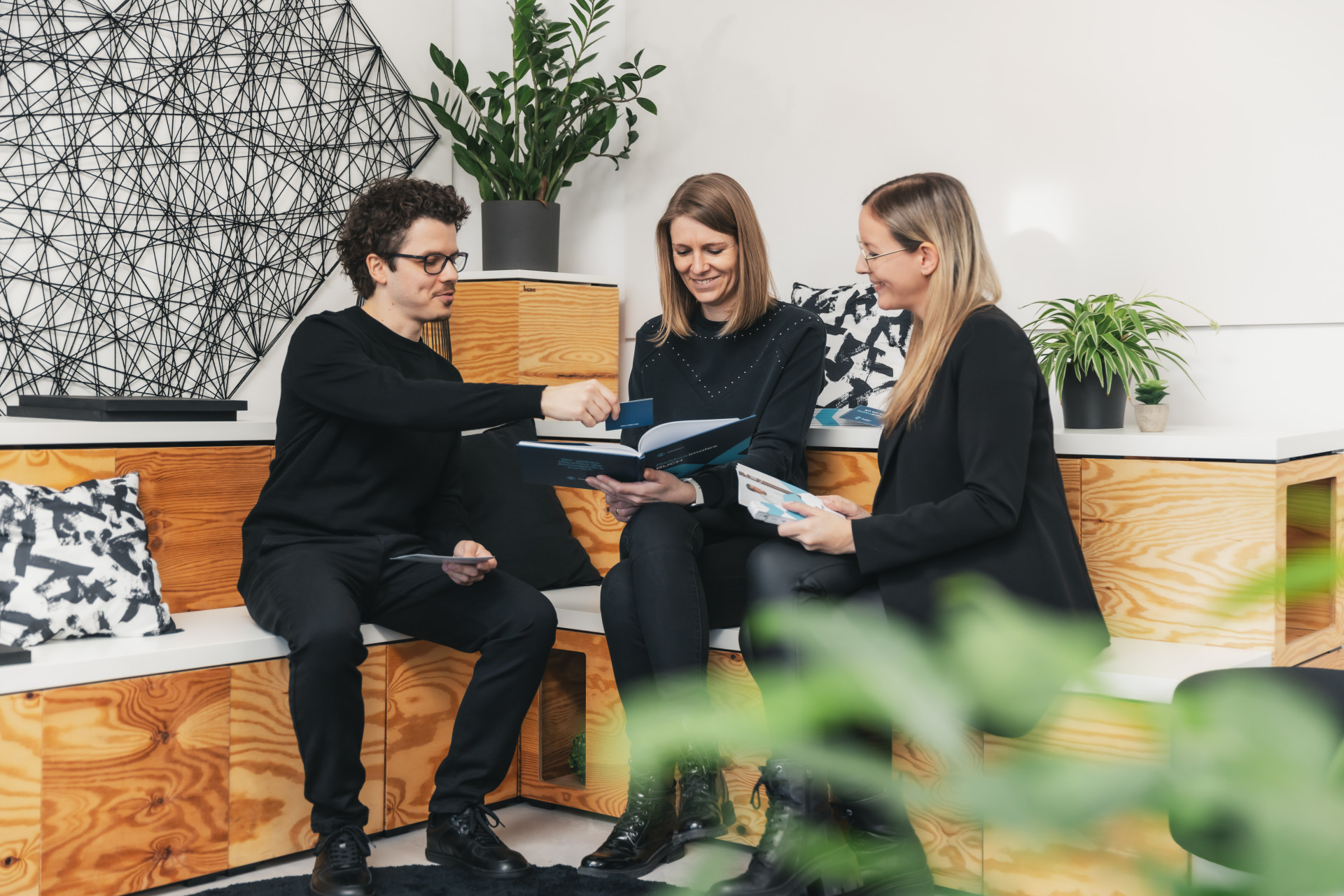 Drei Personen sitzen in einem modernen Büro auf Holzbank. Sie blättern in Zeitschriften. Im Hintergrund sind Pflanzen und eine Wanddekoration aus schwarzen Linien.